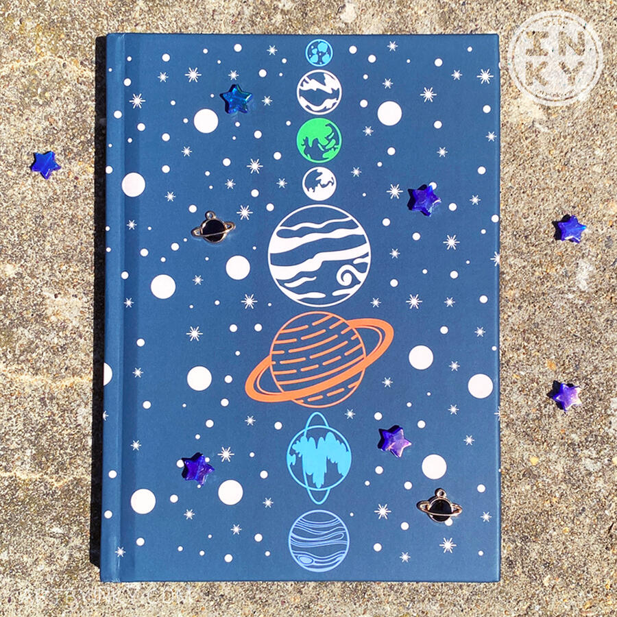 A hardcover blue space journal laid flat frontside up with star and planet charms against a cement background outside.